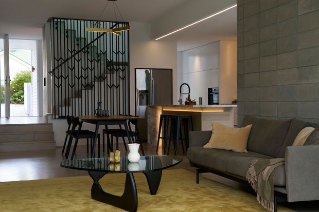 Open-plan contemporary living, dining, and kitchen area with a sculptural glass coffee table and sectional sofa in the foreground. A compact dining table sits beside a dramatic black geometric metal stair screen, while the kitchen features a stone-look island, timber-toned benchtop, integrated appliances, and pendant-style task lighting. Pale timber flooring, a large muted yellow rug, and neutral furnishings create a refined yet warm interior.