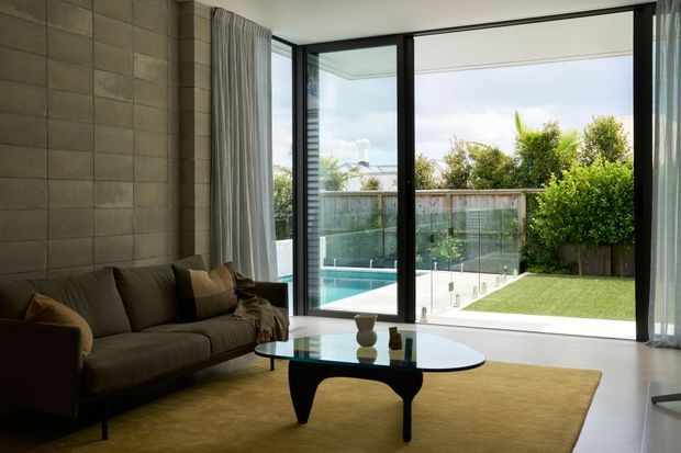 A contemporary living room with a dark upholstered sofa, sculptural glass coffee table, and large warm-toned area rug. A textured block wall forms the feature wall, while full-height black-framed glazing and sliding doors open onto a bright backyard with a swimming pool, lawn, and timber fencing.