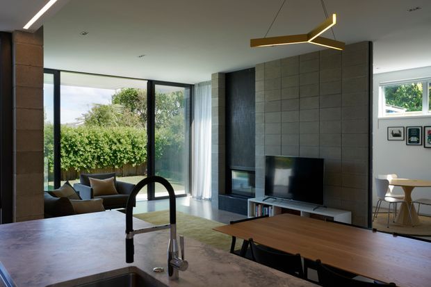 Contemporary open-plan living space viewed from a kitchen island, with a dark tiled fireplace and television wall, floor-to-ceiling glazing opening to greenery, a dining table, and a compact sitting area. The room has a restrained palette of charcoal, grey, timber, and soft neutral furnishings, with linear and sculptural pendant lighting.