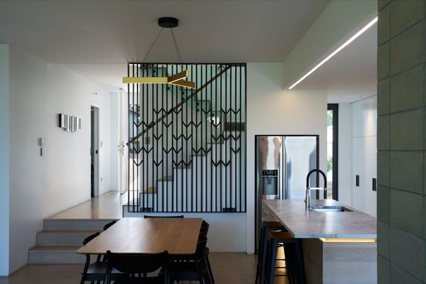 A contemporary open-plan kitchen and dining area viewed from the dining zone. A timber dining table sits in the foreground, while the kitchen features a stone island with an undermount sink, dark tapware, integrated appliances and streamlined cabinetry. A dramatic black metal screen with geometric detailing separates the stairway, and a linear LED strip and sculptural pendant provide architectural lighting. The palette is restrained, combining white walls, warm timber, pale stone and dark metal accents.