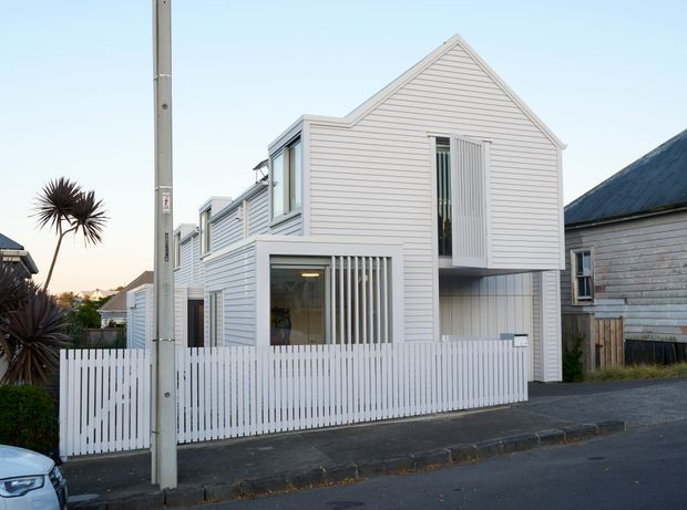 Street-level exterior view of a narrow, multi-level white weatherboard residence. The house has a steep front gable, layered projecting volumes, vertical timber screens, white fencing, and a small upper-level balcony or recessed opening. A neighbouring weatherboard building and suburban street context are visible.