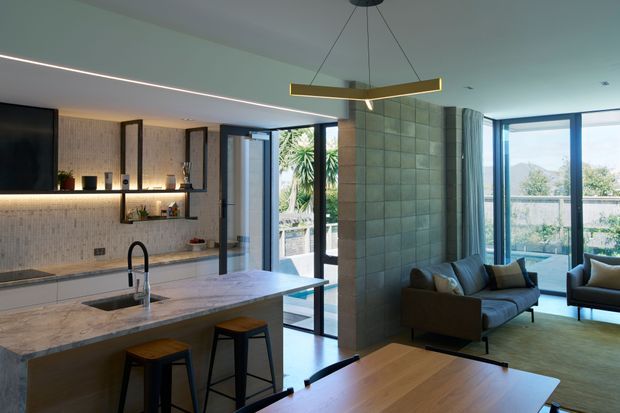 Open-plan contemporary interior combining a kitchen, dining area and living room. The kitchen features a large pale stone island with an undermount sink, black gooseneck tap and two stools, along with pale textured tiled walls, integrated cabinetry and open shelving. A tiled feature partition separates the kitchen from the living area, which has a sofa, armchairs and expansive blue-framed glazing opening toward an outdoor terrace. A linear pendant light, concealed strip lighting and warm timber flooring create a refined, softly lit atmosphere.