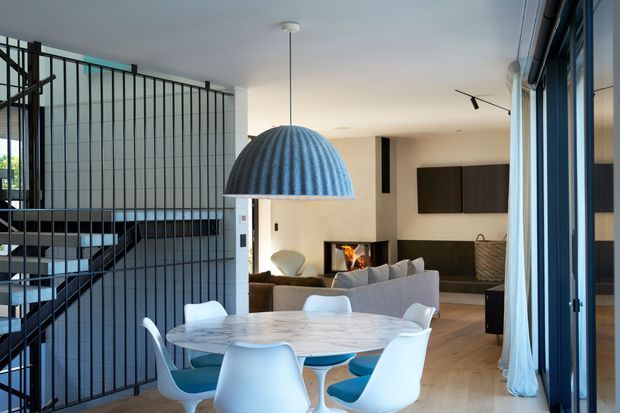 Open-plan contemporary living and dining area viewed from a glazed terrace or balcony. The foreground contains a round marble dining table with modern white and blue chairs, while the background features a light upholstered sofa, fireplace, dark built-in cabinetry, and a compact kitchen zone. A black metal stair balustrade and stairwell dominate the left side, and large glazed openings with curtains line the right. A ribbed blue pendant light hangs over the dining table.