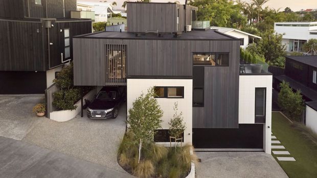 Aerial exterior view of a contemporary multi-level residential home on a sloping suburban site. The house features dark vertical cladding, crisp white rendered wall sections, extensive black-framed glazing, flat roof forms, projecting upper volumes, integrated garages, and landscaped gravel driveways with stepping-stone paths.