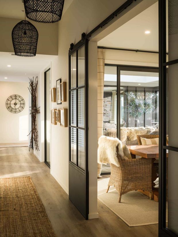 A warm, refined residential interior showing a hallway opening into a dining room through black-framed glazed sliding doors. The hallway has light timber flooring, cream walls, woven runners, framed artwork, a large wall clock, and woven pendant lights. The dining room contains a timber table, wicker dining chairs, textured neutral textiles, and extensive glazing overlooking an outdoor courtyard or garden.