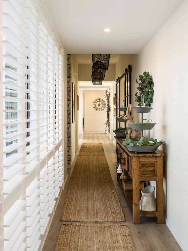 A bright, narrow residential hallway viewed lengthwise toward adjoining rooms. White plantation shutters span the left side, while the right wall features a rustic timber console with layered ceramic planters, greenery, woven decor and small objects. Woven runners extend along the light timber-look floor, and dark woven pendant lights hang in a row overhead. The space is warm, natural and carefully styled with a mix of white walls, timber, rattan and greenery.