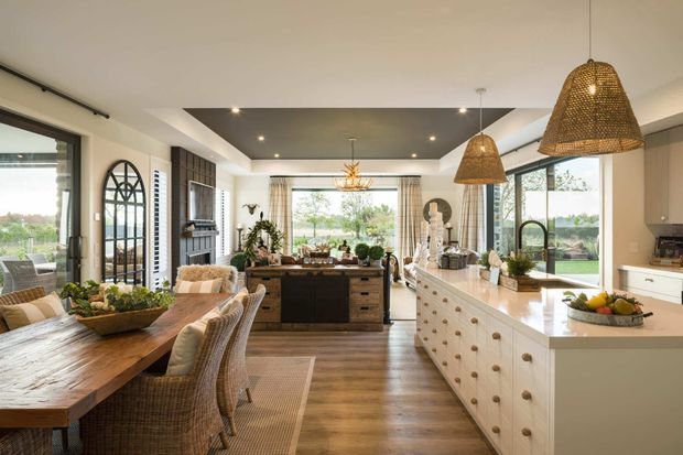 A bright, spacious open-plan kitchen and dining area with a large white island, timber flooring, natural timber cabinetry, woven pendant lights, and a rustic timber dining table with wicker chairs. Full-height glazing and doors open the room to a landscaped exterior, while a dark coffered ceiling creates contrast. The interior has a warm, relaxed contemporary farmhouse character.