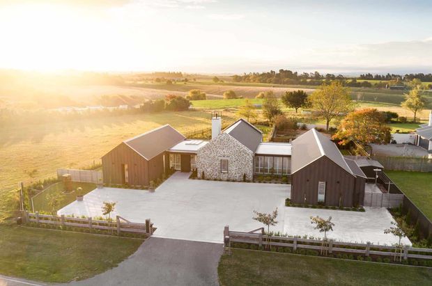 Aerial view of a low-rise rural residential property arranged around a large pale concrete courtyard. The home combines dark timber-clad pitched-roof wings with a central light stone-clad section, skylights, a chimney, and extensive glazing. Fenced lawns, a driveway, and expansive cultivated fields and trees surround the house, with warm low-angle sunlight creating a tranquil countryside setting.