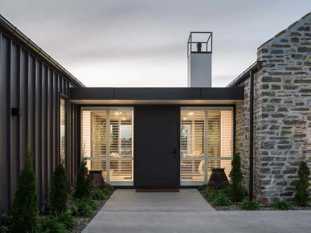Front exterior view of a compact contemporary building addition or entry pavilion, framed between a dark vertically ribbed metal-clad wall on the left and an existing rough stone masonry wall on the right. The entrance has a flat dark roof, black central door, and large sidelights with timber-toned horizontal blinds. A tall rectangular chimney or flue rises above the roof, and a simple concrete path leads through planted beds to the entry.
