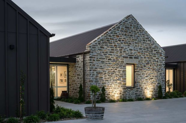 Exterior view of a modern rural-style building complex with a prominent gabled stone masonry facade, dark metal-clad wings, and warm illuminated windows. The courtyard is finished with smooth concrete paving and restrained planted landscaping under an overcast sky.