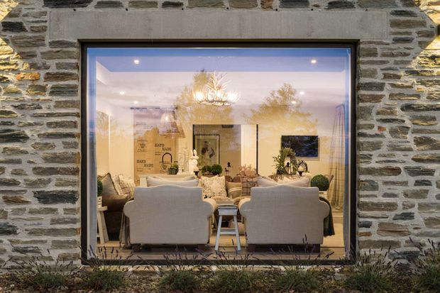 Exterior view through a large glazed opening into a warmly lit traditional living room. The building facade is constructed from irregular grey natural stone with light mortar joints and a dark window frame. Inside, two large light upholstered armchairs face into the room, with additional seating, a central coffee table, decorative plants, artwork, and an ornate chandelier visible. The scene has a refined, cosy evening atmosphere.