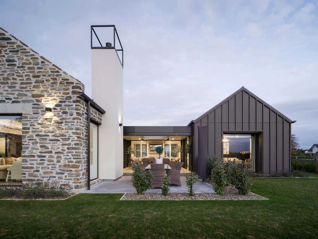 Exterior view of a contemporary residential composition combining a renovated stone structure with a dark vertical-panel gabled wing. A tall white chimney-like architectural element with a minimalist black frame rises between the volumes. Large glazed openings connect the interior living areas to a paved outdoor dining or lounge terrace, surrounded by lawn and low planting.