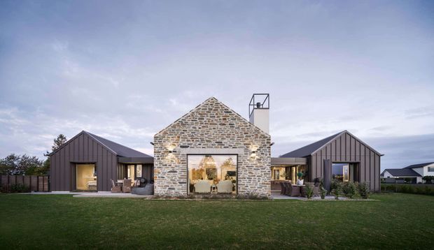 A low, symmetrical residential composition with a central gabled stone-clad pavilion and large glazed opening, flanked by two dark vertically clad gabled wings. The central volume has a simple chimney, while the wings feature dark metal or timber-style cladding and large windows. A broad lawn occupies the foreground, with neighbouring homes and an overcast sky in the background. The overall architecture is contemporary with a strong farmhouse-inspired form.