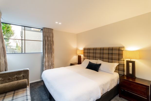 A warm, softly lit bedroom with a large upholstered plaid feature headboard, neatly made bed, dark bedside tables, table lamps, patterned curtains, carpeted floor, and a large black-framed window. The neutral beige and cream palette creates a refined, inviting atmosphere.