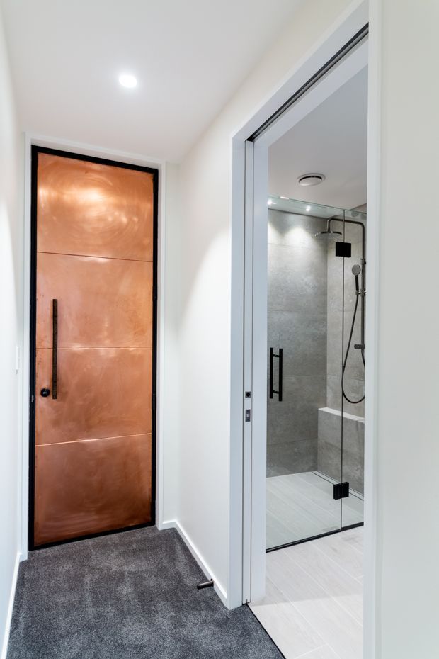 A contemporary hallway or entry area with dark grey carpet, white walls and trim, a distinctive copper-toned metal door with a black vertical handle, and an open bathroom doorway. The bathroom contains a frameless glass shower enclosure with black hardware, grey textured wall tiles, pale floor tiles, and a built-in bench.