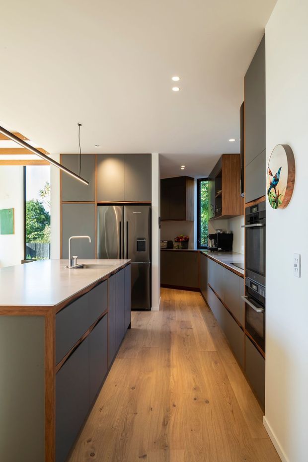 A contemporary galley-style kitchen with a large island, dark matte cabinetry edged in warm timber, stainless-steel refrigerator, integrated wall ovens, white stone benchtops, and warm natural timber flooring. The space has a clean open-plan feel, recessed ceiling lights, a linear pendant over the island, and views to greenery through the windows.