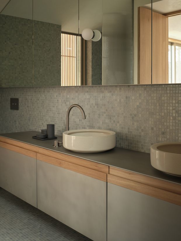 Contemporary bathroom vanity with twin vessel basins, a dark stone-look benchtop, warm timber trim, pale grey-green cabinetry, and small-format grey mosaic wall and floor tiles. A large mirrored wall reflects the room, while ribbed glass and timber screening add texture and privacy. The palette is muted, earthy, and refined.