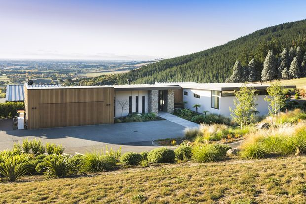 A low, single-storey contemporary home set into a sloping landscaped site, viewed from above and across the driveway. The house has a broad flat roof, white rendered walls, dark vertical facade elements, a stone-clad entry section, and a large timber-toned garage door. Layered native-style planting and ornamental grasses surround the residence, with an expansive valley and densely forested hillside beyond.