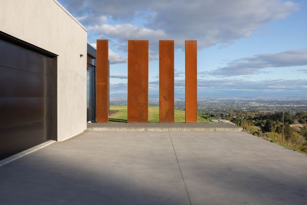 Exterior view of a contemporary residence on an elevated site, showing a broad concrete driveway or terrace, a dark garage door, pale rendered wall, and a row of tall rusted steel feature screens. A wide urban and landscape vista is visible beyond the property under a partly cloudy sky.