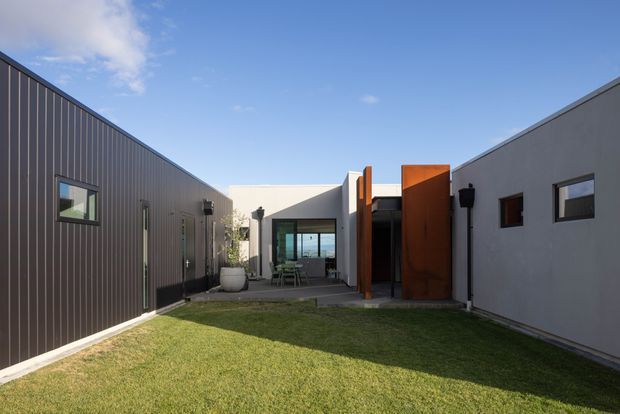 A contemporary single-storey courtyard-style building viewed from a landscaped grass courtyard. The composition shows dark vertical cladding on the left wing, a light rendered or smooth-finished wing on the right, and a central glazed opening with a striking rust-coloured metal screen or entry feature. Clean geometric forms, simple rectangular windows, and strong contrasts define the architecture.