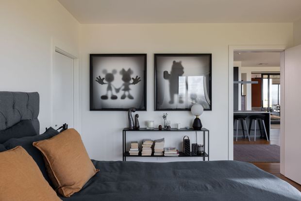 A contemporary bedroom with a dark upholstered bed, charcoal bedding, and warm tan cushions. A slim black console with books and decorative objects sits against a white wall beneath two framed monochrome artworks. An open doorway reveals a connected kitchen or dining area with dark cabinetry, island seating, and timber flooring.