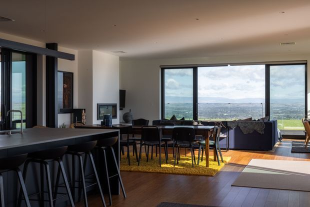 Open-plan contemporary living and dining area with a large dark kitchen island and bar stools in the foreground. A dining table and chairs sit beside a sofa and television, while expansive floor-to-ceiling glazing frames a broad landscape view. Warm timber flooring, dark window frames, neutral walls, and abundant daylight create a refined, spacious interior.