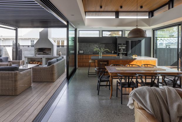 Open-plan contemporary kitchen and dining area with a timber island, dark cabinetry, stone or terrazzo-style flooring, and a large dining table. Full-height glazing opens to a covered outdoor lounge and fireplace area, while a timber-lined ceiling, skylight, pendant light, and black-framed glazing add architectural definition.