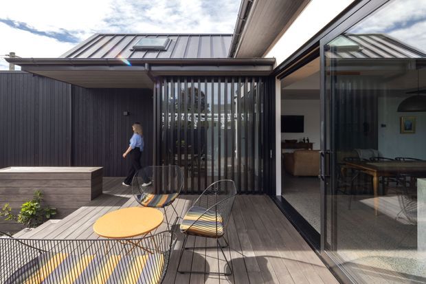 A contemporary residential courtyard and timber deck viewed from outdoors. The composition features dark vertical cladding, a metal pitched roof, a covered outdoor seating area with a slatted privacy screen, and large glazed sliding doors opening to a dining and living space. Wire-frame chairs and a round orange table occupy the deck, with a raised timber planter at left.