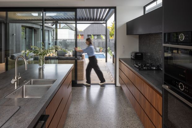 Contemporary open-plan kitchen with a large central island, double stainless-steel sink and curved tap, dark stone benchtops, timber cabinetry, integrated black appliances, and a textured dark splashback. Full-height glazing opens toward a covered outdoor terrace and courtyard, bringing in natural light and creating a strong indoor-outdoor connection.