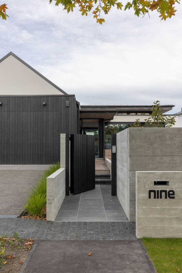 A contemporary residential entrance viewed from the street, featuring a dark vertical-clad facade, a black pedestrian gate, grey concrete boundary walls, large-format charcoal paving, and a covered entry beyond. The minimalist house has a simple gabled roof form alongside a low horizontal entry canopy, with landscaped grasses and small trees framing the approach. A wall-mounted sign reads “nine”.