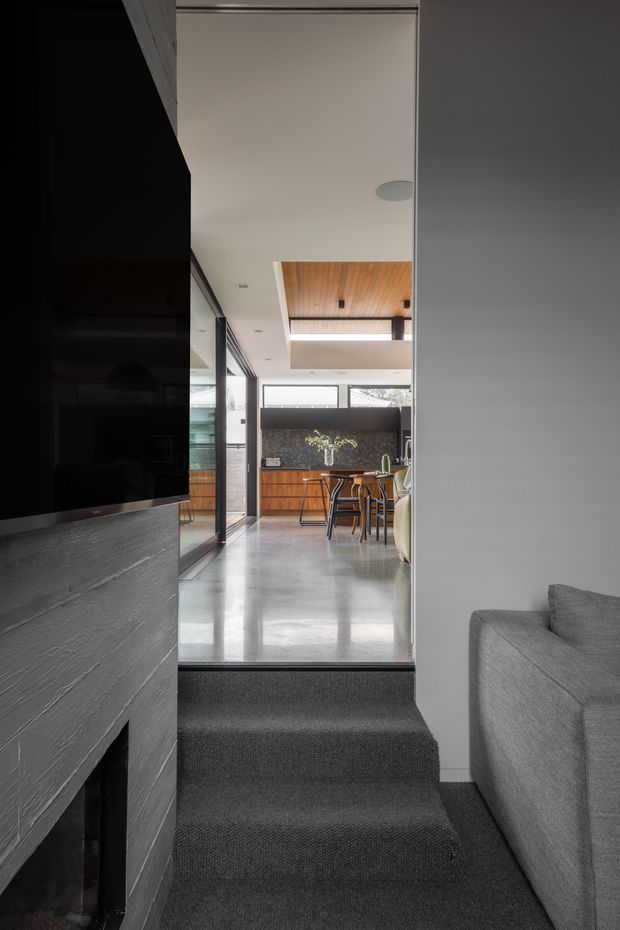 A contemporary open-plan interior viewed from a carpeted living room, looking up two steps into a bright dining and kitchen area. A large dark television and textured dark fireplace surround occupy the left foreground, with a grey sofa on the right. The adjoining space has polished concrete flooring, timber kitchen cabinetry, a dark stone or tile splashback, timber ceiling lining, high-level windows, and a dining table with chairs. The palette is predominantly charcoal, grey, white, and warm timber, with strong linear architectural detailing and a partial ceiling void.