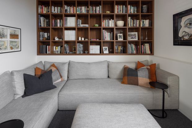 A cozy living room with a large light-grey L-shaped sectional sofa and matching ottoman, accented by rust, charcoal, and beige cushions. A full-width dark timber built-in bookcase occupies the main wall, displaying books and decorative objects. White walls, dark carpet, framed artwork, and a small black side table complete the refined, modern interior.