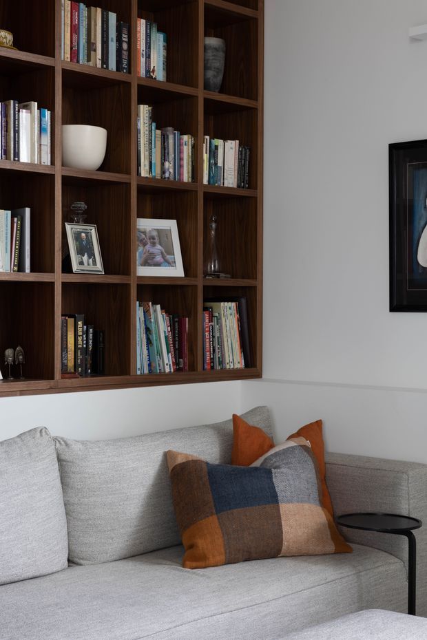 Close-up interior view of a refined living room corner featuring a large dark timber built-in bookshelf filled with books and decorative objects, a light grey upholstered sofa, and layered rust, navy, and brown cushions. A black-framed artwork and slim black side table are visible against white walls. The space has a warm, contemporary and tailored feel.