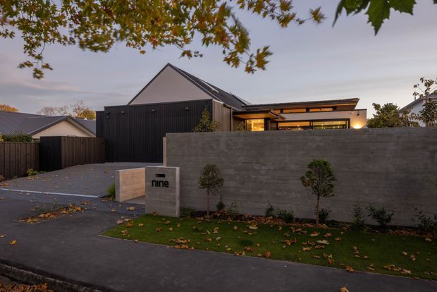 Street-facing exterior view of a contemporary residential home at dusk. The property has a dark vertical-clad main volume with a steep gabled roof, a lower flat-roofed wing with warm interior lighting, a tall smooth concrete boundary wall, dark paved driveway, and a landscaped lawn with clipped shrubs. A concrete entry monument bears the text “nine”.