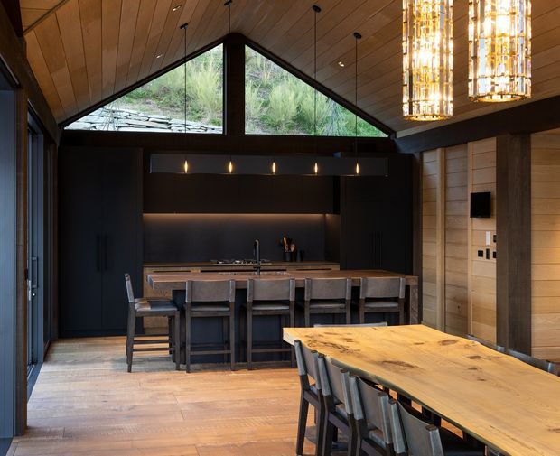 A warm, dramatic open-plan kitchen and dining space with a steep vaulted timber ceiling and high triangular clerestory glazing overlooking vegetation. The kitchen features matte-black full-height cabinetry, a timber island with integrated sink and seating for several people, dark structural beams, and subtle under-cabinet lighting. A large timber dining table with dark chairs occupies the foreground, while a tall sculptural illuminated pendant hangs above. Timber wall panelling and dark-framed glazing reinforce the lodge-like character.