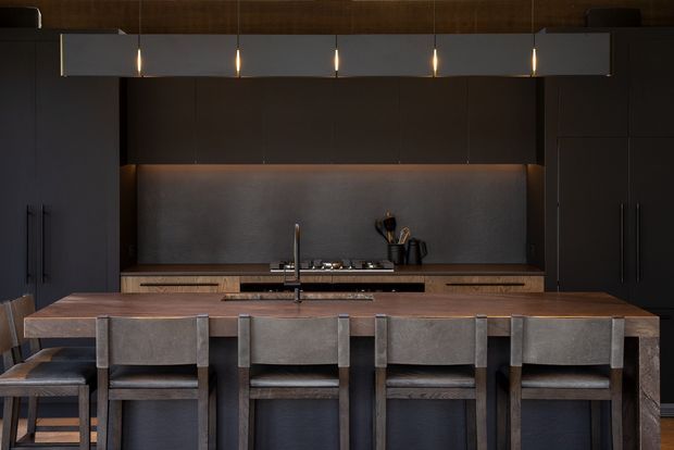 A dark, sophisticated contemporary kitchen with a long timber island and four upholstered bar stools in the foreground. Full-height dark cabinetry frames a concealed cooking and preparation zone, with a metallic grey splashback, timber cabinetry, integrated cooktop, sink and tap, and warm linear pendant lighting. The palette is moody and neutral with rich timber accents and subtle warm illumination.