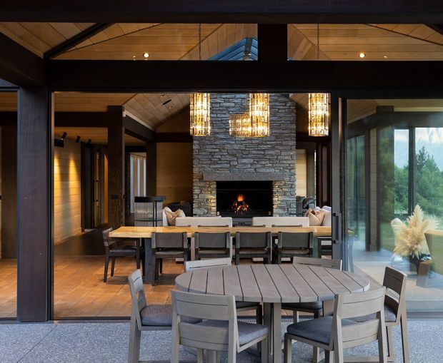 View from an outdoor terrace into a sophisticated open-plan living and dining space through large glazed sliding doors. The interior features a dramatic stacked-stone fireplace wall, exposed dark timber structural framing, warm timber ceiling and flooring, sculptural pendant lights, and a dining table aligned with a lounge area. A separate round outdoor dining table and chairs occupy the foreground.
