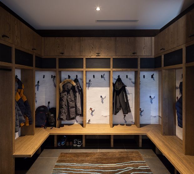 A custom timber-lined mudroom or ski storage room with built-in open lockers, upper cabinets, benches, hooks, and cubbies. Coats, ski clothing, boots, and bags are stored throughout the joinery. The locker back panels feature snowy mountain-themed imagery, while dark trim, warm wood, and subdued lighting create a practical, rustic alpine atmosphere.