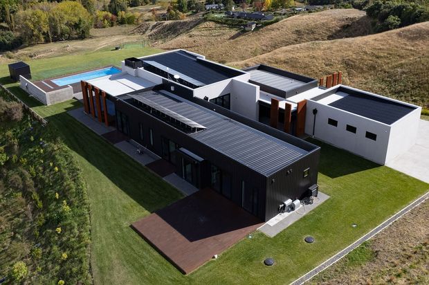 Aerial view of a large contemporary single-storey residence arranged as several connected rectangular volumes. The building has dark corrugated metal-clad sections, white rendered walls, flat or very low-pitched dark roofs, timber-toned vertical screening, extensive dark-framed glazing, and landscaped lawns. A rectangular swimming pool and paved outdoor area are visible at the rear, with rolling rural terrain surrounding the property.