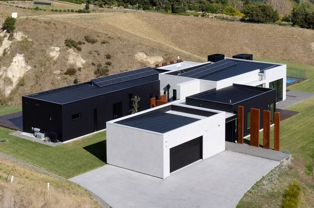 Aerial view of a contemporary multi-wing residential property set into a landscaped, sloping rural site. The building has crisp white and black rectangular volumes, flat roofs, extensive rooftop solar panels, dark cladding, large garage doors, and timber-toned vertical privacy screens. Paved driveways and neatly maintained lawns surround the house.