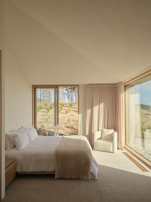 A serene, softly lit bedroom with a minimalist arrangement. The room features a large upholstered bed with white bedding and a textured taupe throw, a cream armchair, full-height glazed windows and sliding doors, and floor-length pleated curtains. Warm neutral finishes and expansive views toward a dry, landscaped outdoor setting create an airy, calm atmosphere.
