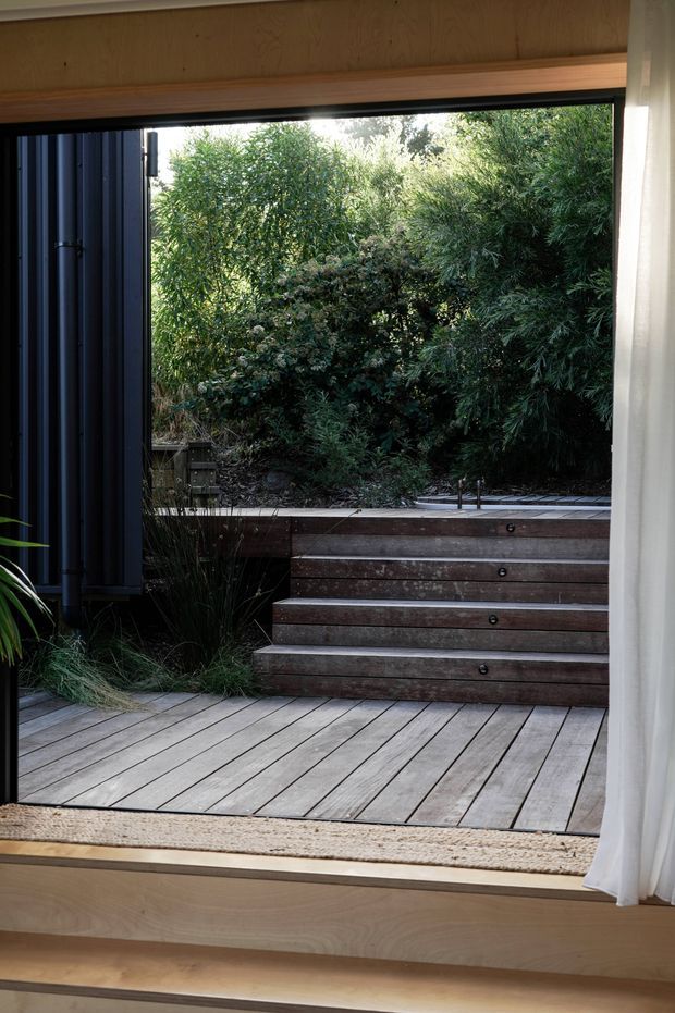 View from inside through a large opening onto a compact timber deck and a short set of broad timber steps leading into dense landscaped planting. The interior edge has light timber wall and sill detailing, with dark vertical exterior cladding at the left and a sheer curtain at the right. The scene feels connected to a private garden and uses natural timber and dark cladding in a contemporary architectural composition.