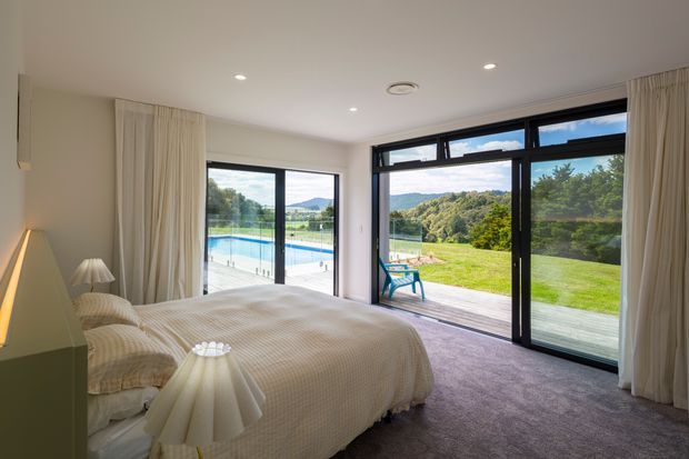 Bright, softly styled bedroom with a large upholstered bed, cream bedding, bedside lamps, full-height curtains, and dark-framed glazing. The glazing opens toward a timber deck, lawn, swimming pool, and leafy hills beyond.