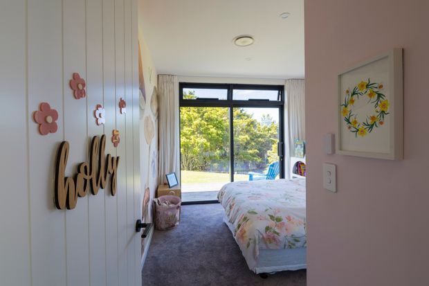 A bright, cosy bedroom viewed from the doorway, with a floral-patterned bed, grey carpet, pale pink walls, and a white vertical-panel feature wall decorated with playful flower ornaments and a wooden “hello” sign. Full-height glazing at the far end opens the room to leafy outdoor views, with curtains framing the window. A framed floral artwork and light switches are visible on the right wall.