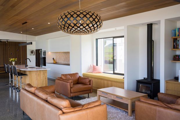 Open-plan contemporary living, dining and kitchen area with warm timber ceilings and wall detailing, white cabinetry, a stone kitchen island, tan leather sofas and armchairs, and a black freestanding wood-burning stove. A large woven pendant light, built-in window seat, shelving and expansive glazing add warmth and texture.