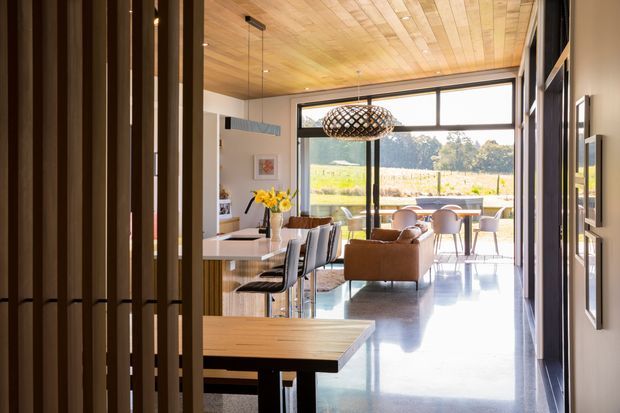 A warm, contemporary open-plan interior viewed from a hallway or entry zone, looking toward a combined dining and living area. The space features a timber-lined ceiling, polished concrete flooring, dark-framed full-height glazing, timber dining furniture, upholstered seating, and a sculptural woven pendant light. A rural landscape is visible beyond the glazing.