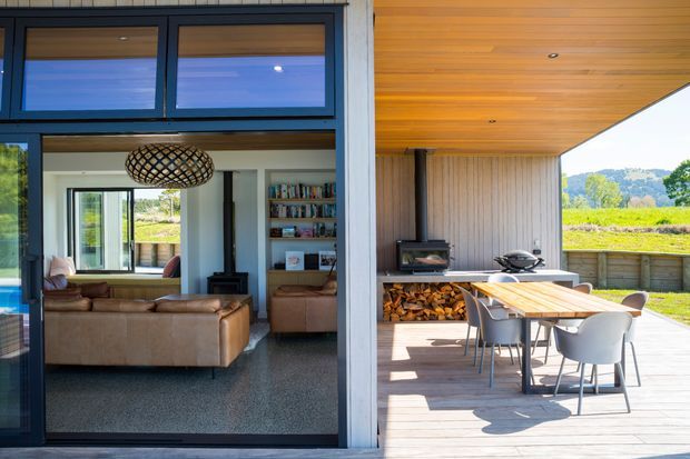 A bright contemporary indoor-outdoor living space viewed from the deck. Large dark-framed sliding glass doors open to a living room with leather sofas, a patterned pendant light, shelving and a wood-burning stove. The adjoining covered outdoor area has a timber-lined ceiling, vertical timber wall cladding, a built-in fireplace with stacked firewood, a barbecue, and a dining table with chairs. Rural landscape is visible beyond the deck.