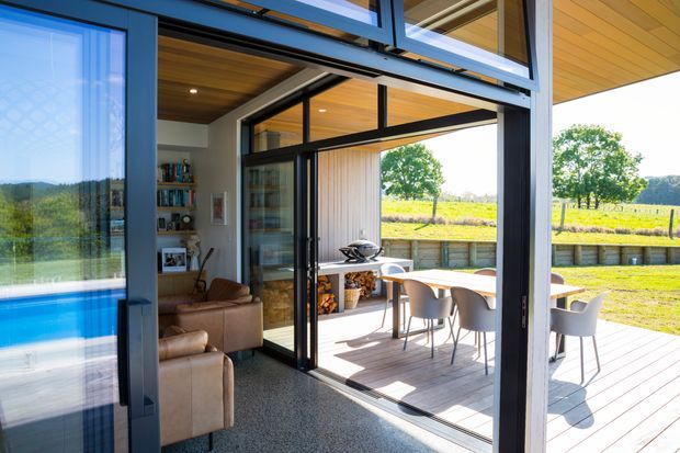 A bright contemporary indoor-outdoor living area opening onto a timber deck and rural landscape. The image shows large black-framed glazed sliding or folding doors, a warm timber-lined ceiling, a lounge with leather seating and shelving, and an outdoor dining setting with a built-in barbecue area. A swimming pool is visible through the glazing on the left.