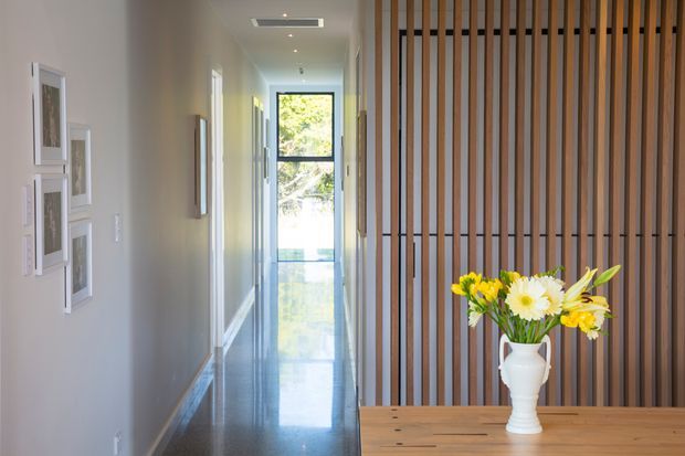 A bright, narrow contemporary hallway viewed from an entry or living area. The corridor has light neutral walls, framed artwork, polished dark flooring, recessed ceiling lighting, and a full-height glazed door or window at the far end. A timber slat screen partially divides the hallway from a timber console table topped with a white vase and yellow-and-white flowers.