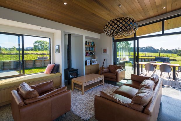 Warm, light-filled contemporary living room with a timber-lined ceiling, white walls, expansive black-framed glazing, and views onto a green rural landscape. The room is furnished with multiple brown leather sofas and armchairs around a timber coffee table, with a built-in window seat, open shelving, a compact black wood-burning stove, and a sculptural woven pendant light. Glazing opens to an outdoor dining terrace.